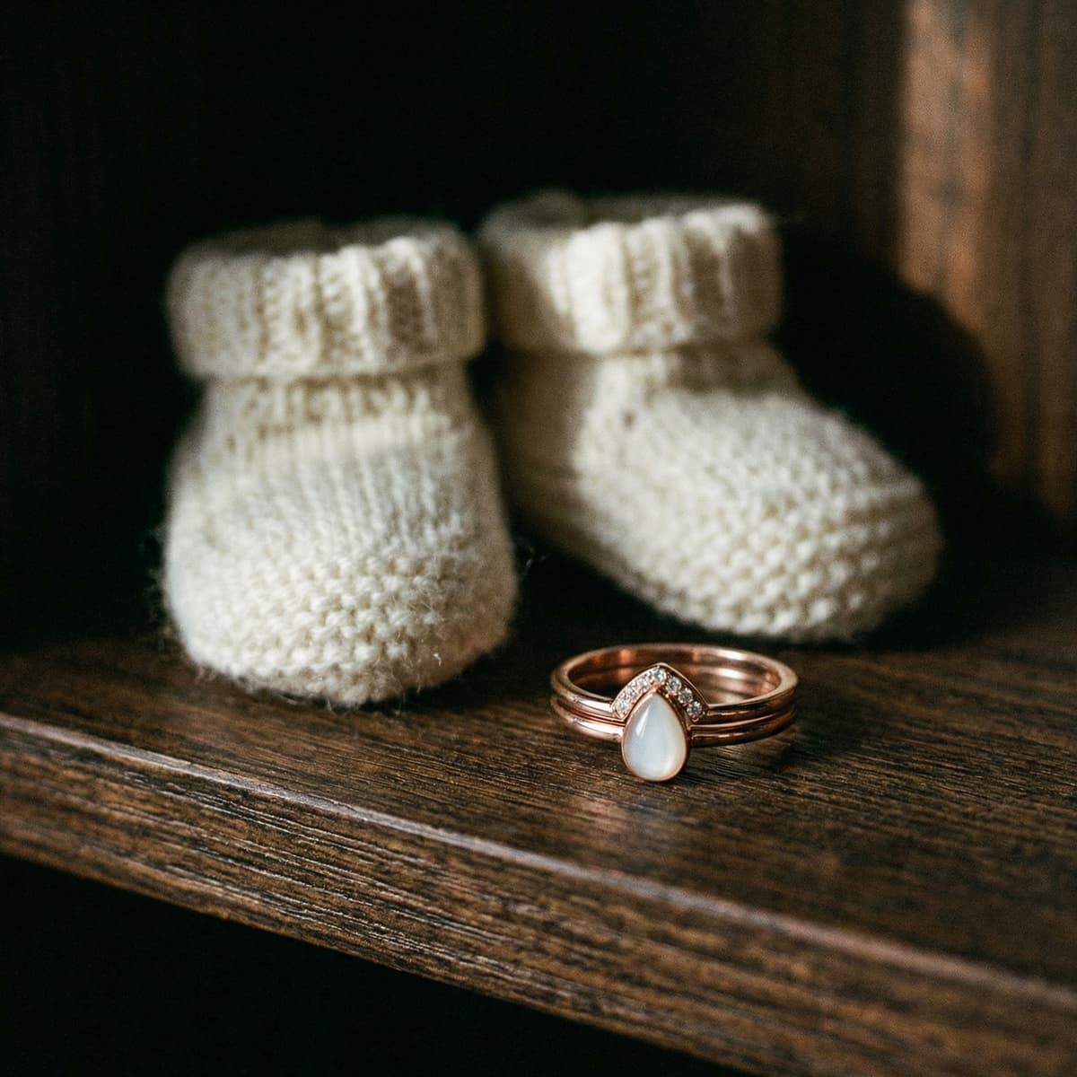 White knitted baby booties on a rustic wooden shelf beside a rose-gold ring with a teardrop breastmilk stone, a sentimental baby keepsake for mom