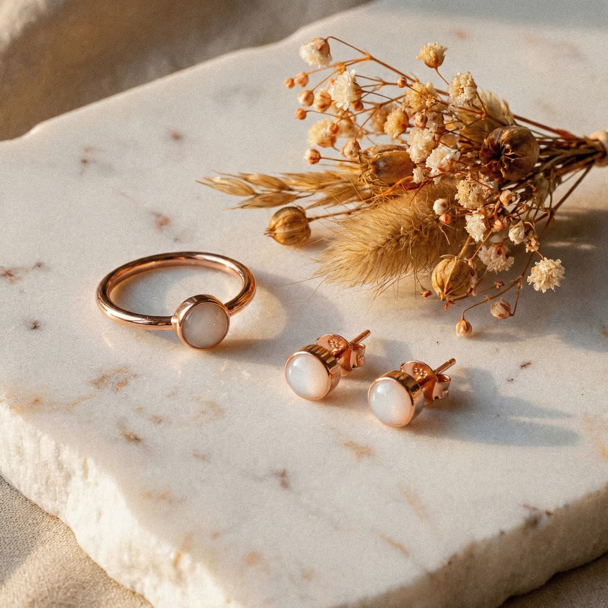 Rose-gold breastmilk jewelry set with milky white resin stone ring and matching stud earrings on a marble slab beside dried flowers, a sentimental choice among the best push presents for new moms.