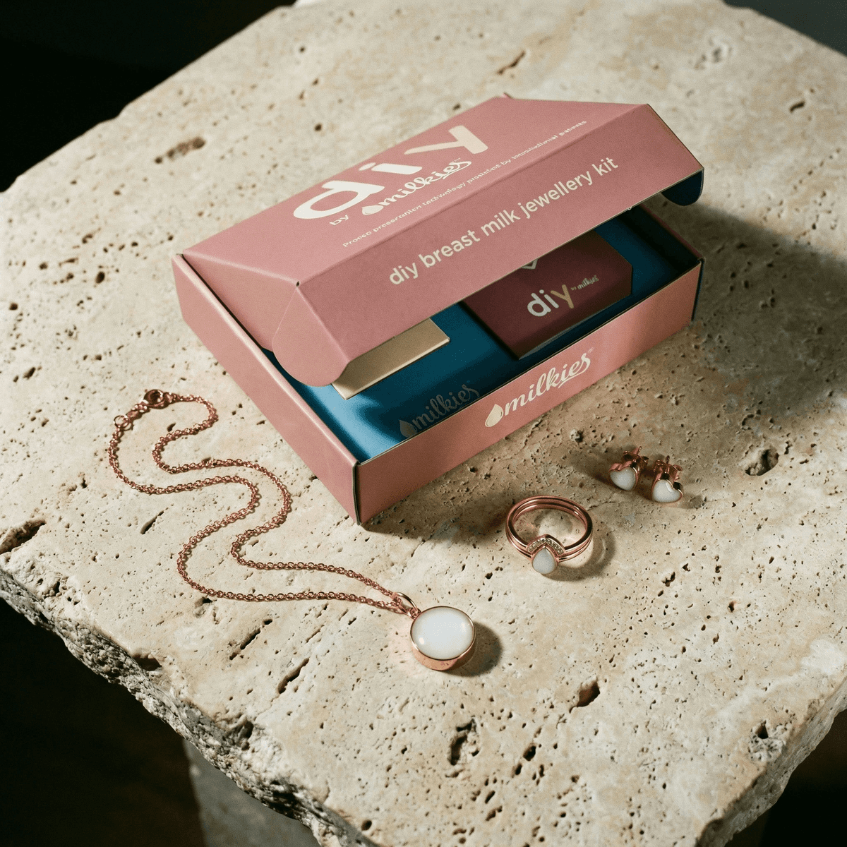 Open DIY by Milkies breast milk jewellery diy kit box on a stone surface with a rose-gold necklace, ring, and stud earrings featuring white keepsake resin stones.