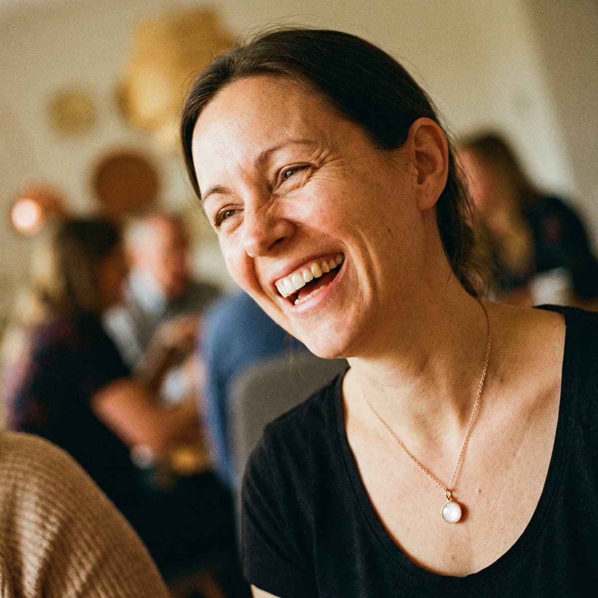 Smiling mother in a softly blurred indoor setting wearing a milky-white breastmilk stone pendant necklace during a warm breastfeeding celebration moment