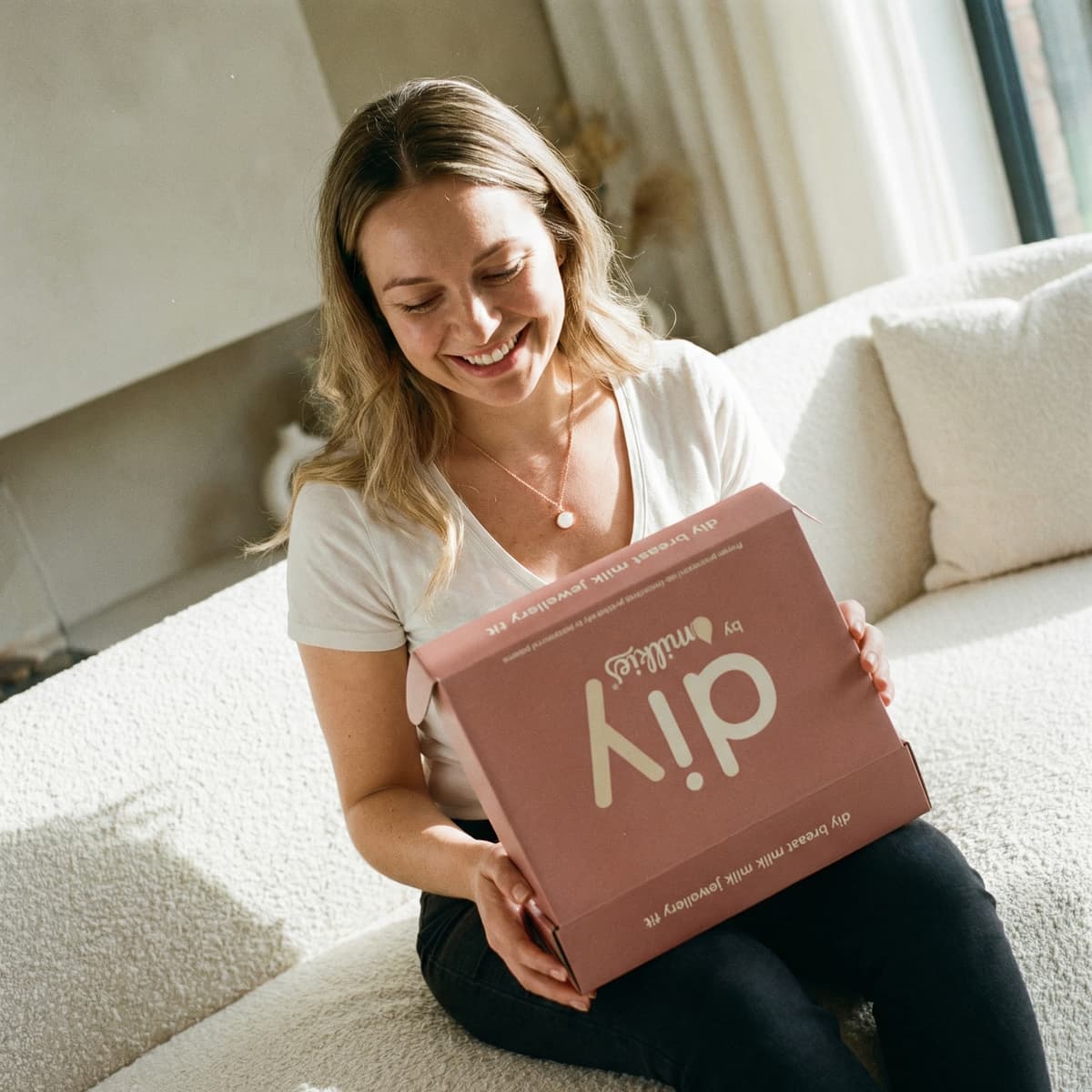 Smiling new mom sitting on a sofa holding a DIY by MILKIES breastmilk jewelry kit box, a thoughtful breastfeeding new mother gift that highlights why making a personal keepsake at home is worth it.