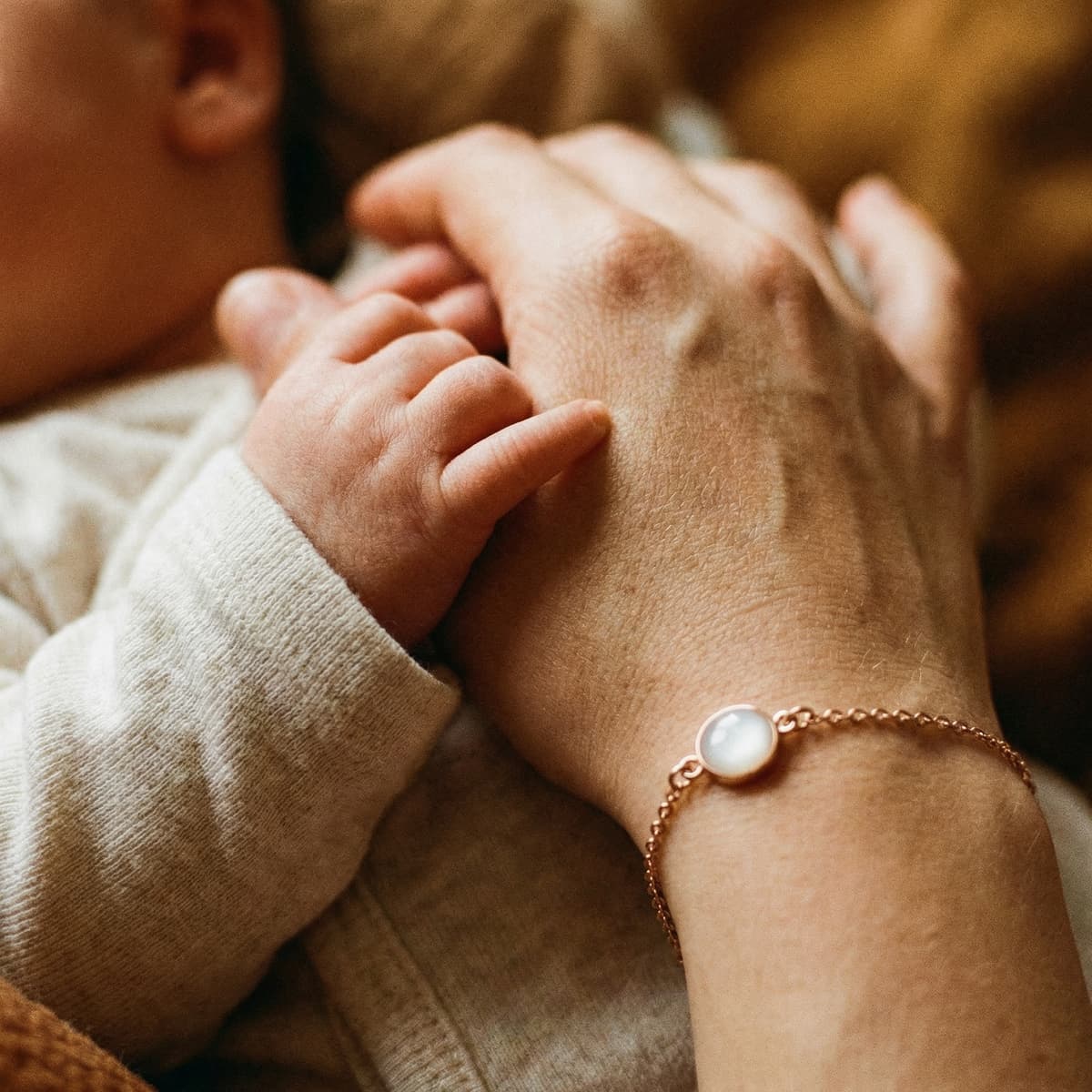 Mother holding a newborn’s hand while wearing a delicate rose-gold keepsake bracelet made with preserved breastmilk in a milky white resin stone close-up