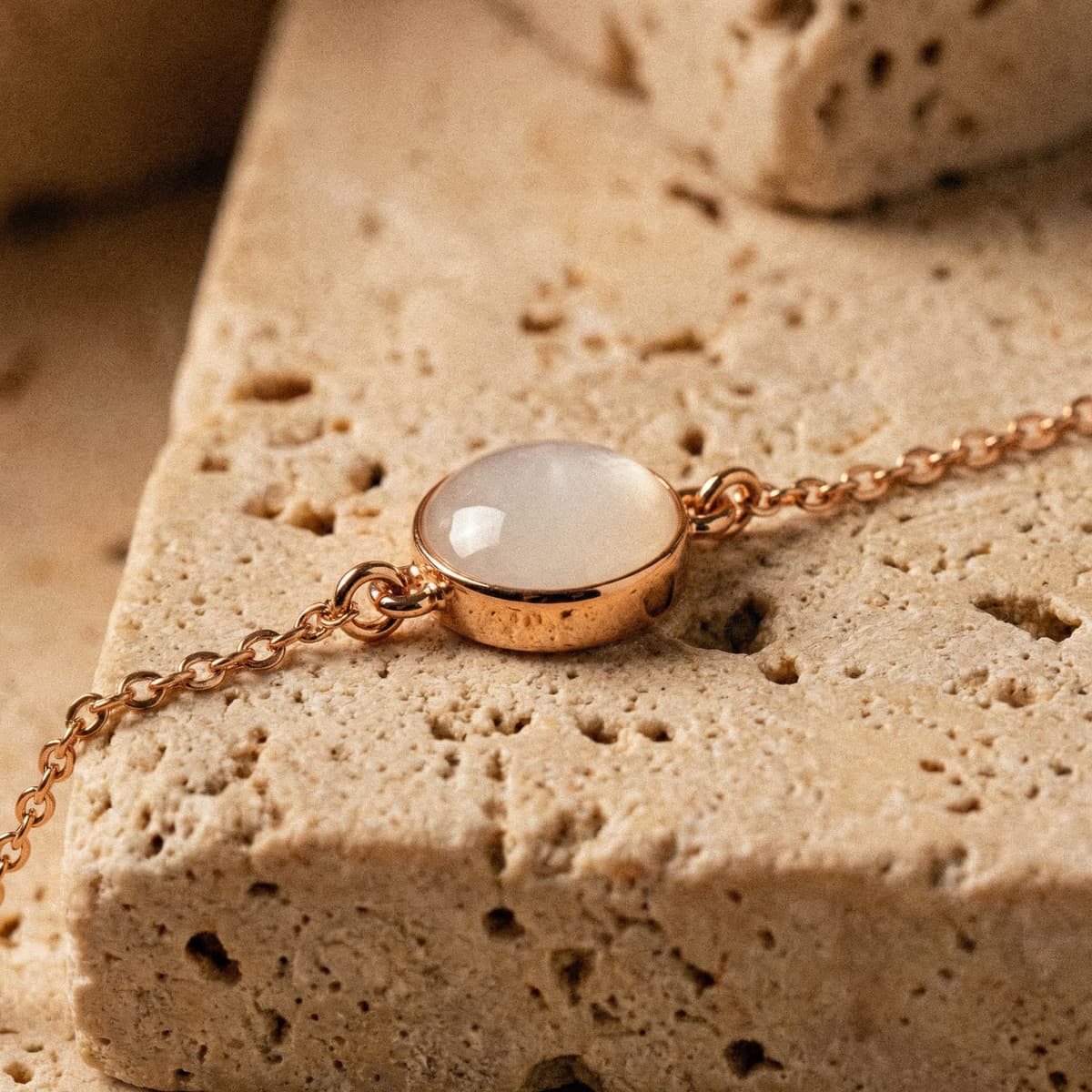 Rose-gold chain bracelet with a milky white resin cabochon charm, a delicate breastmilk bracelet displayed on a textured beige stone background.