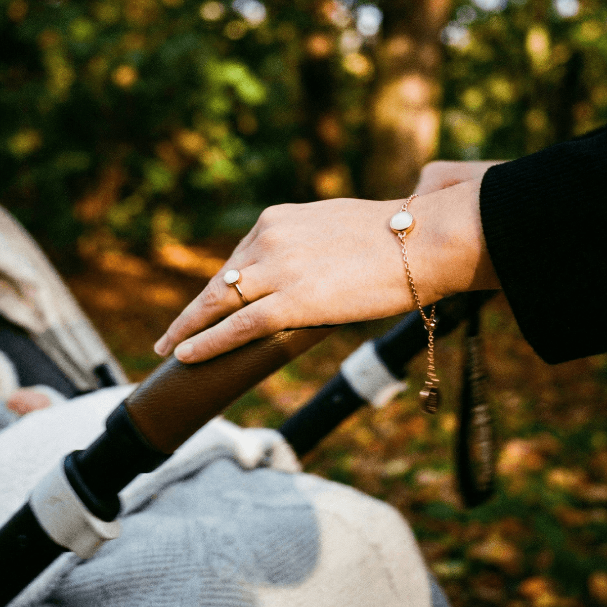 Mother’s hand pushing a stroller outdoors, wearing a delicate gold chain bracelet with a milky-white resin charm made from a breastmilk bracelet diy kit keepsake jewelry set.