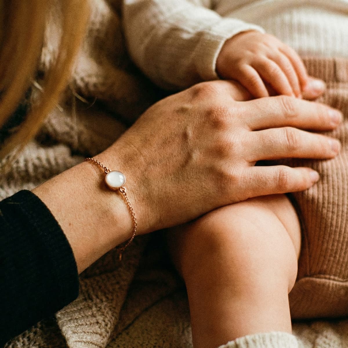 Close-up of a mother holding her baby’s hands while wearing a delicate rose-gold keepsake bracelet with a milky white resin stone from a breastmilk bracelet kit.
