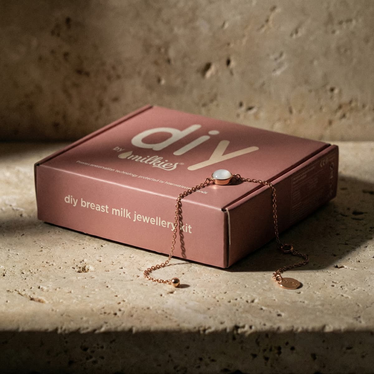 Blush pink DIY by Milkies box for an at-home breastmilk bracelet making kit, featuring a rose-gold chain bracelet and white resin stone on a textured beige surface