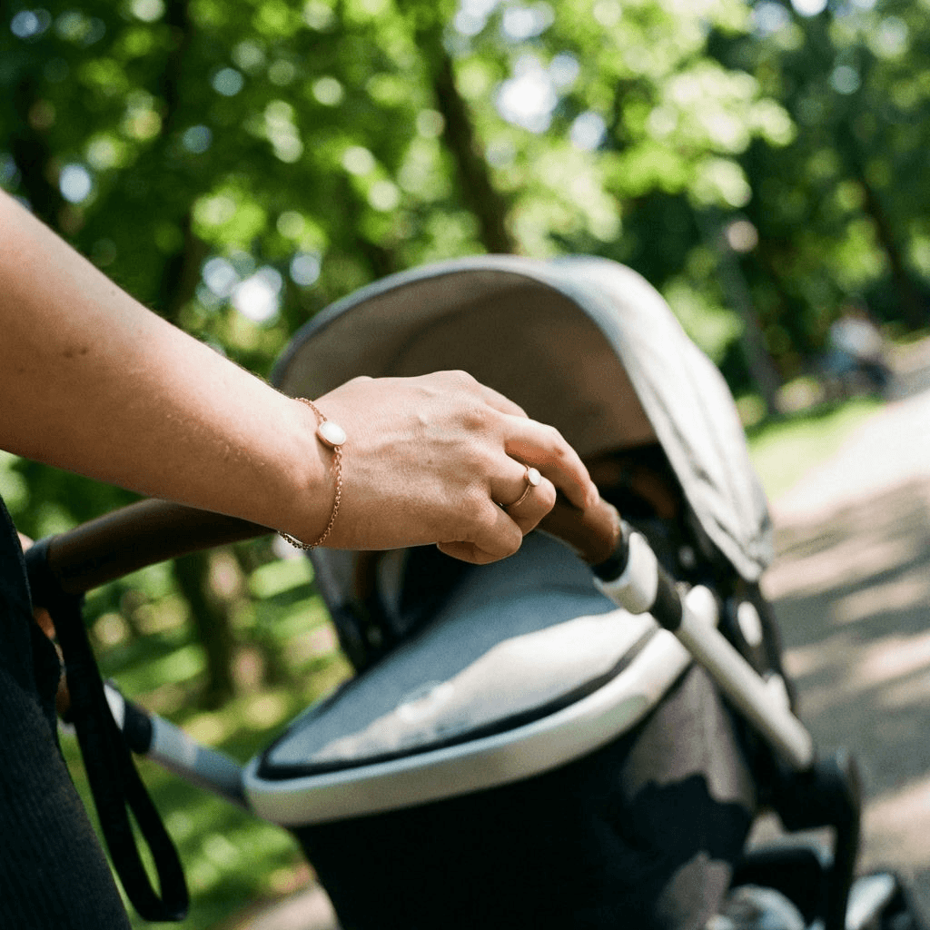Mother’s hand pushing a baby stroller outdoors while wearing a delicate gold bracelet with a milky-white stone made using a breastmilk bracelets kit.