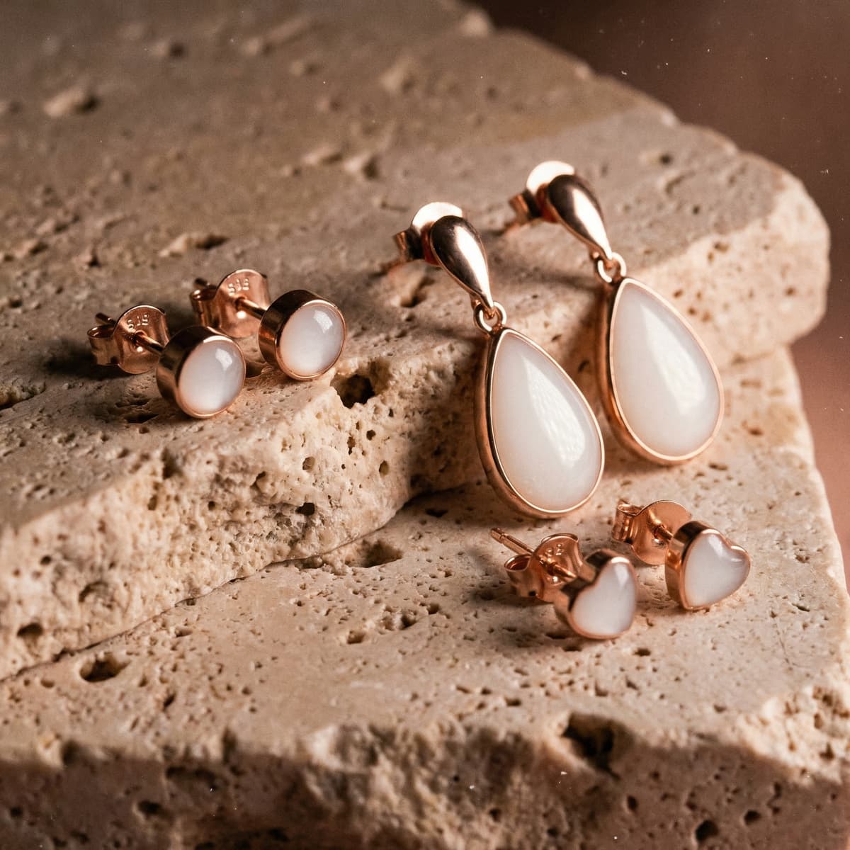 Rose gold jewelry set featuring a breastmilk earring design with milky white resin teardrop drops, round stud earrings, and heart studs displayed on a textured stone surface.