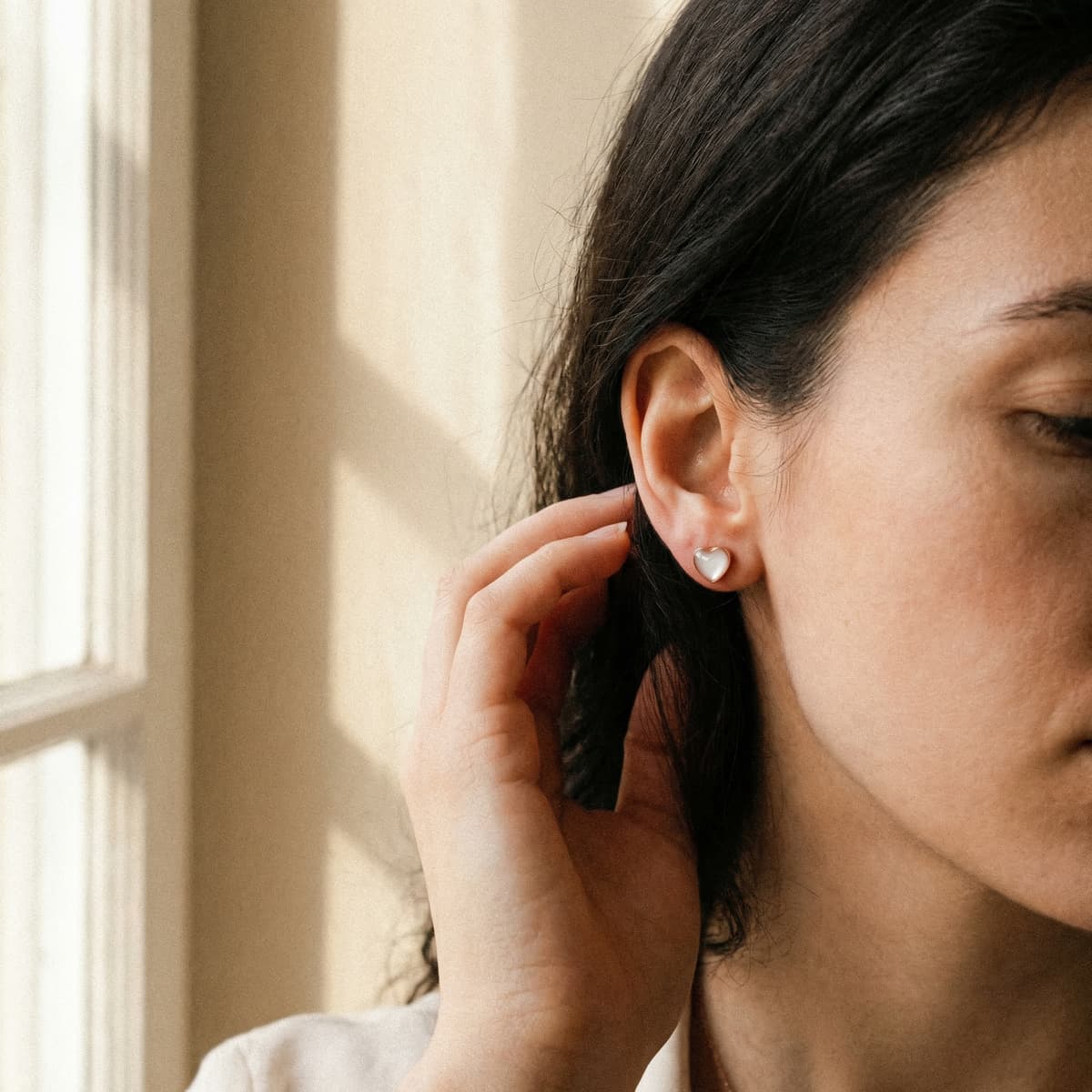 Woman wearing small heart-shaped stud earrings made with a breastmilk earring kit, photographed in soft natural window light.