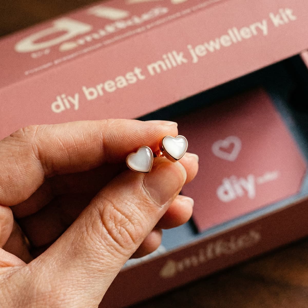Hand holding heart-shaped resin breastmilk earrings in front of the DIY by MILKIES breast milk jewellery kit box.