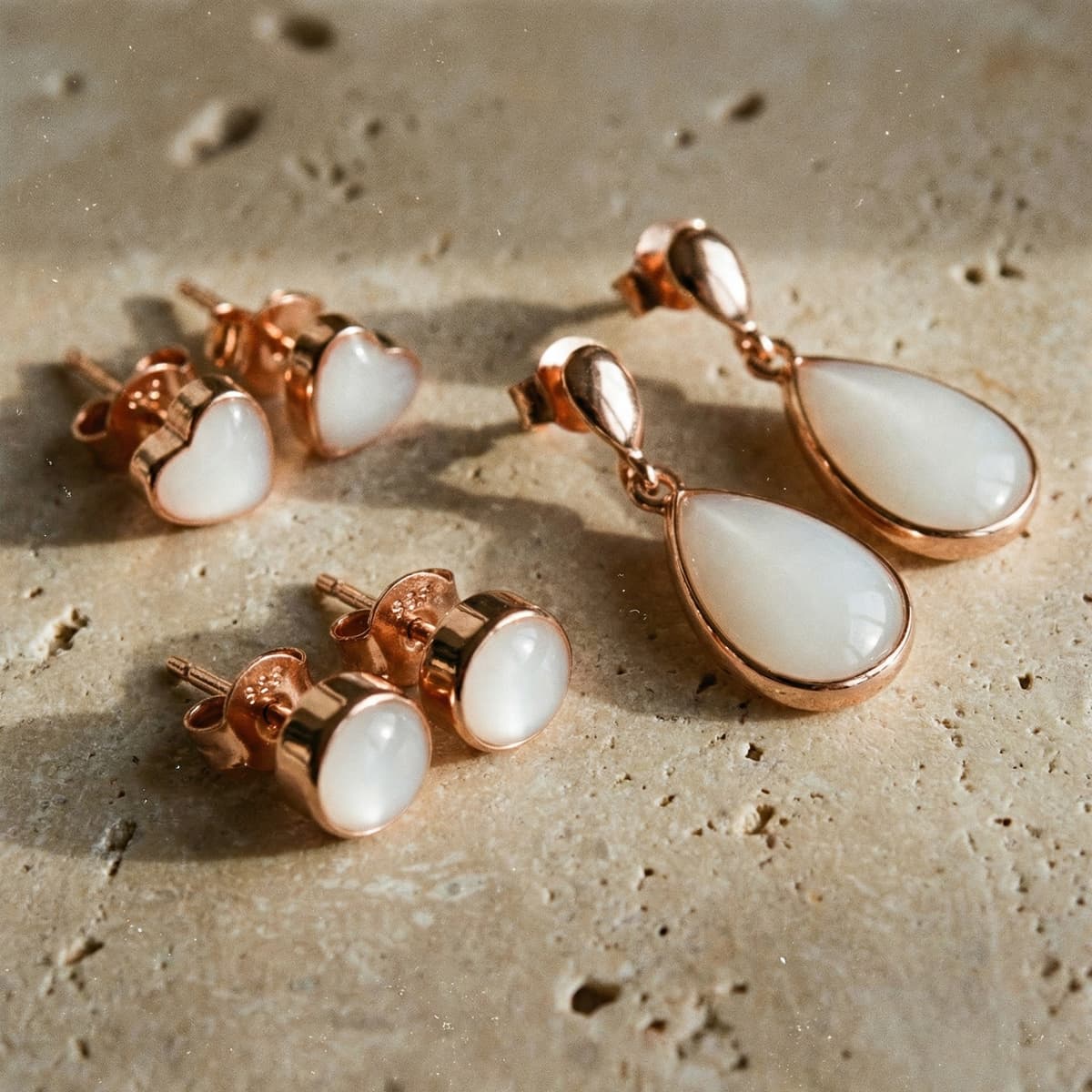 Rose gold jewelry set with breastmilk earrings, including heart studs, round studs, and teardrop drop earrings with milky white resin stones on a neutral stone background