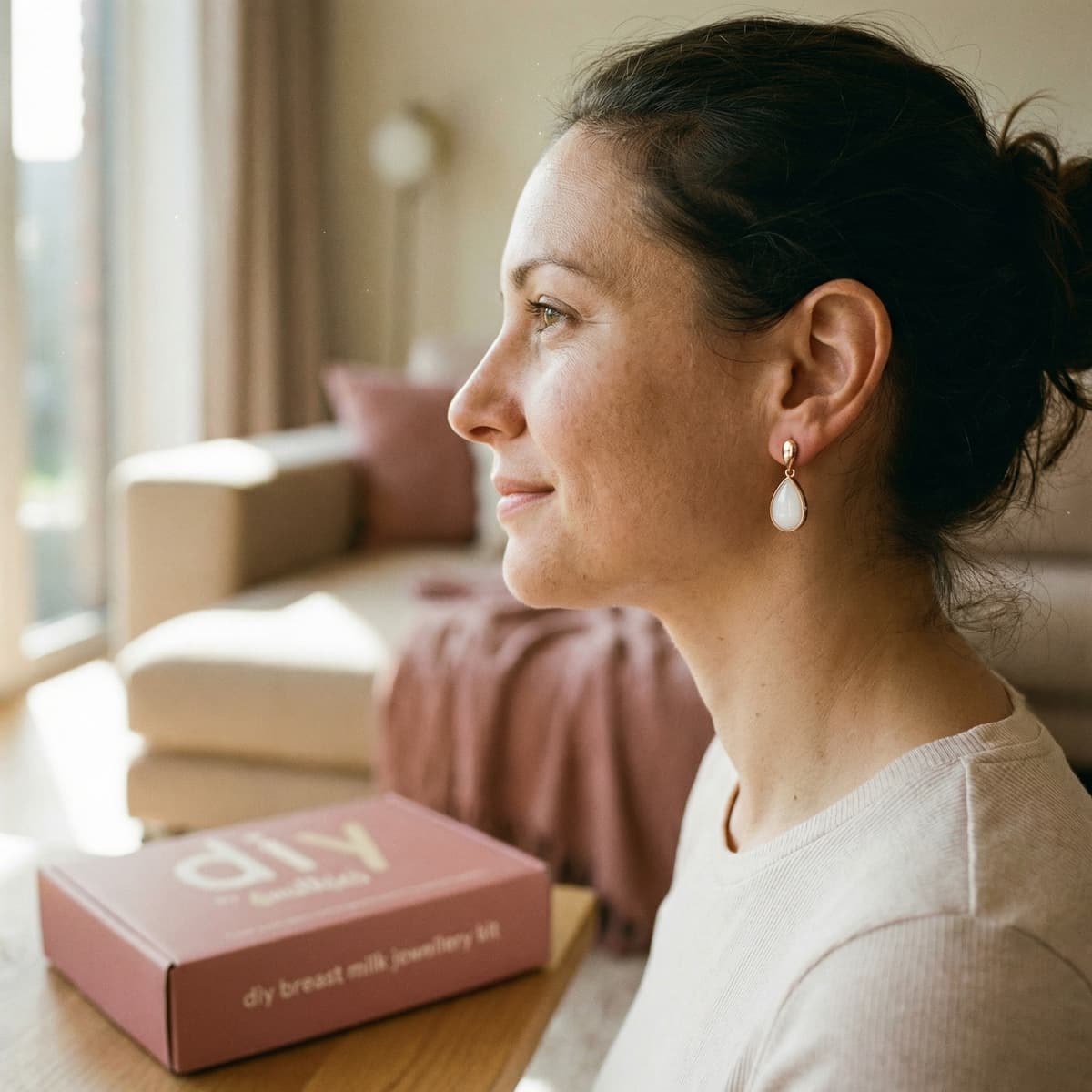 Woman wearing delicate breastmilk earrings at home beside the DIY by MILKIES breast milk jewellery kit box, highlighting why making a personal keepsake yourself is a meaningful choice.