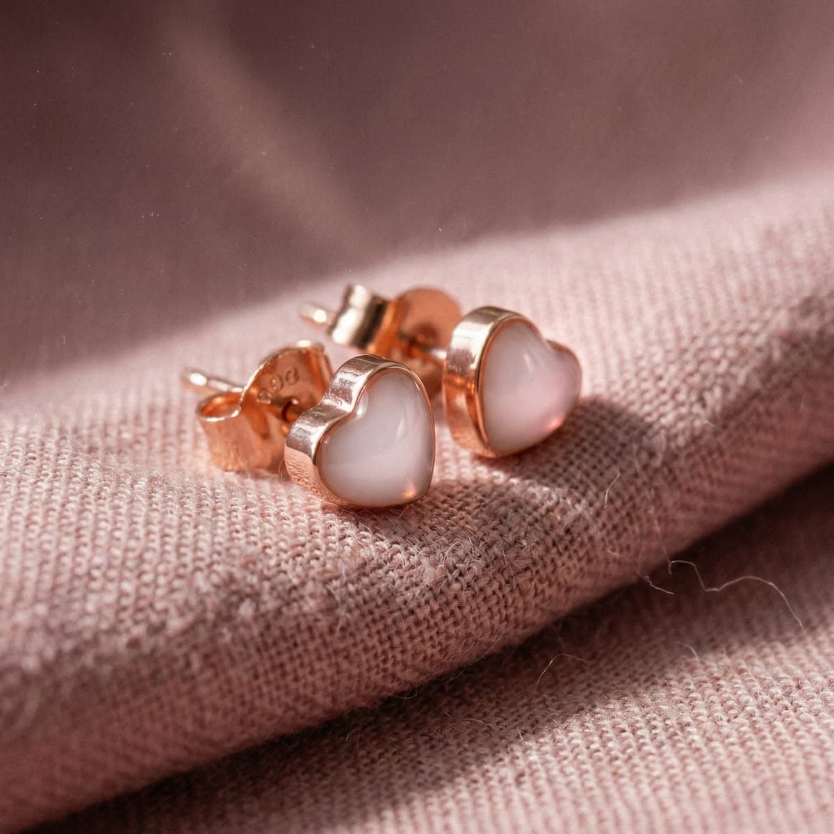 Rose gold heart-shaped stud earrings with milky white resin shown close-up on soft pink fabric, inspired by a breastmilk earrings kit keepsake jewelry design.