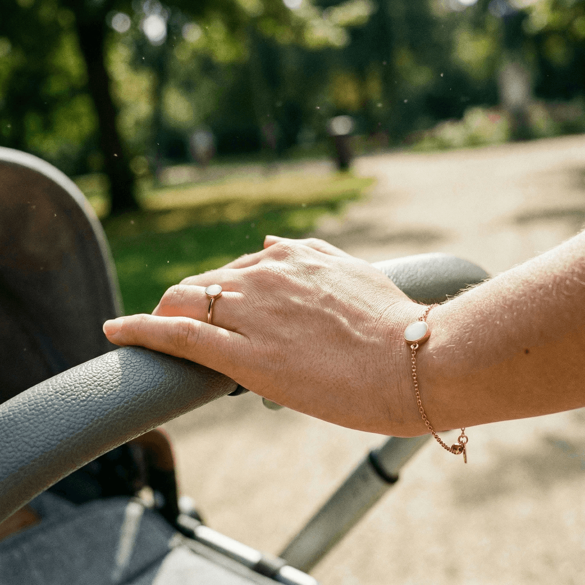 Hand resting on a stroller handle outdoors, wearing minimalist breastmilk jewellery—a gold ring and matching bracelet with oval white milk-stone settings.
