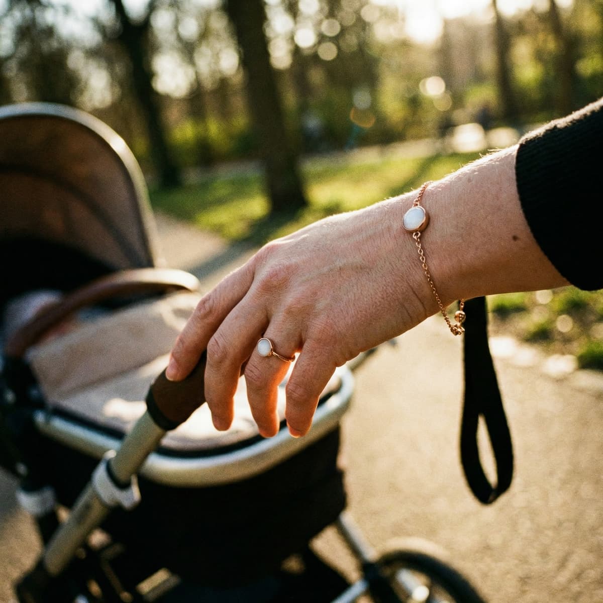 Mother’s hand pushing a stroller outdoors in warm sunlight, wearing a gold ring and bracelet with milky white stones as a subtle breastmilk keepsake jewelry set.