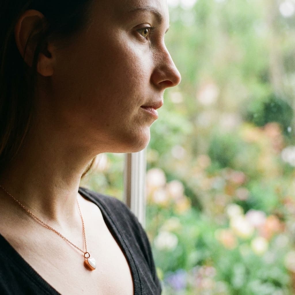 Mother wearing a minimalist gold pendant made with a breastmilk necklace diy kit, standing by a window and showcasing a delicate resin keepsake necklace