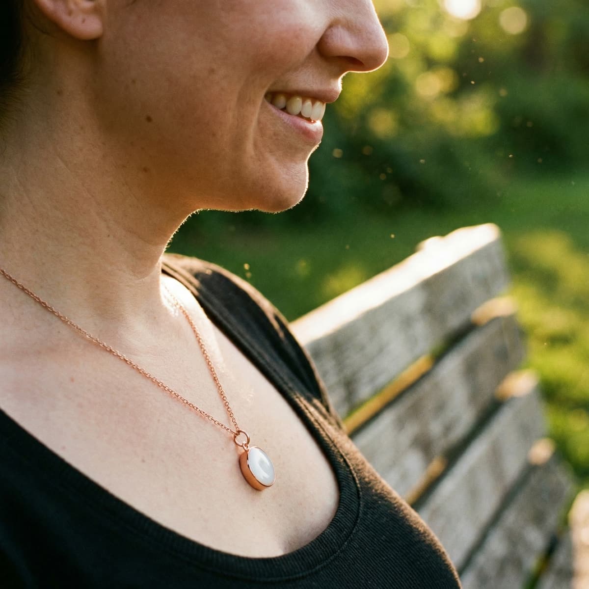 Close-up of a smiling woman outdoors wearing a rose-gold oval keepsake pendant on a delicate chain, made using a breastmilk necklace kit.