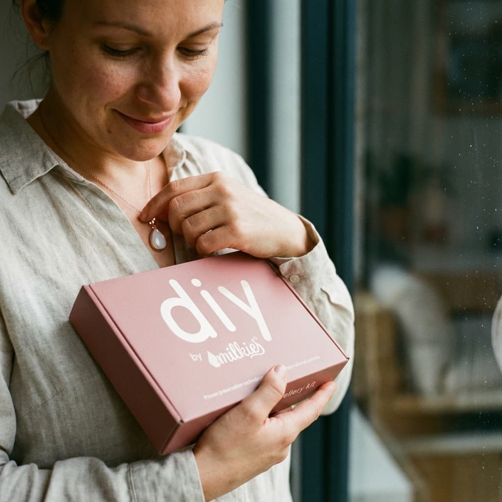 Mother holds a DIY by MILKIES box and touches her keepsake pendant, showing why an at-home breastmilk necklace kit is a meaningful, simple way to create a personal memory at home.