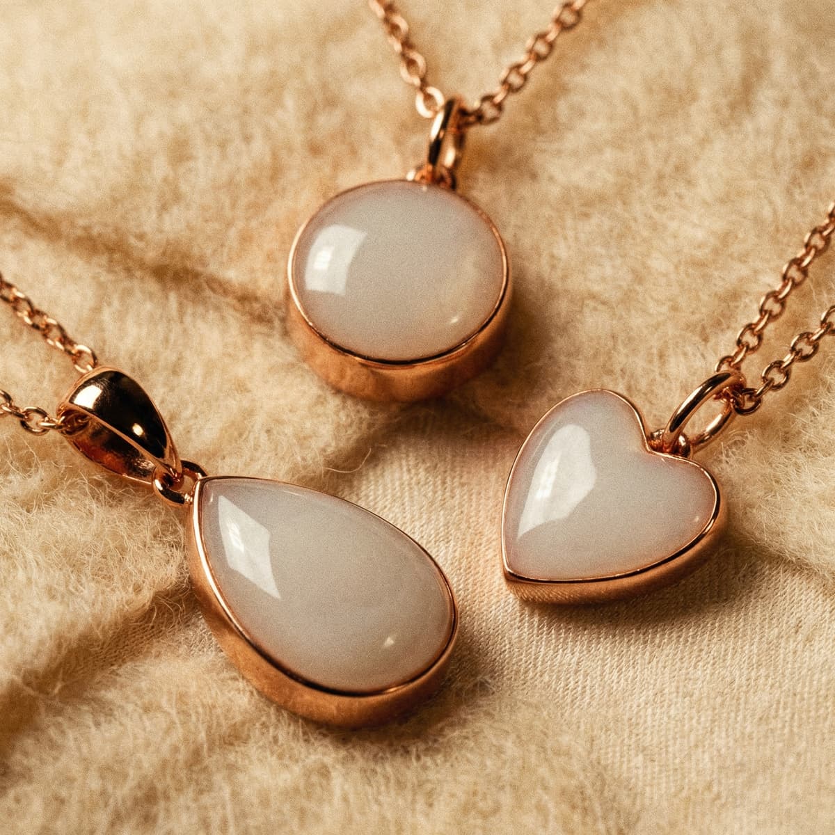 Close-up of three rose-gold breastmilk necklaces with creamy white resin stones in round, teardrop, and heart-shaped pendants on soft beige fabric background