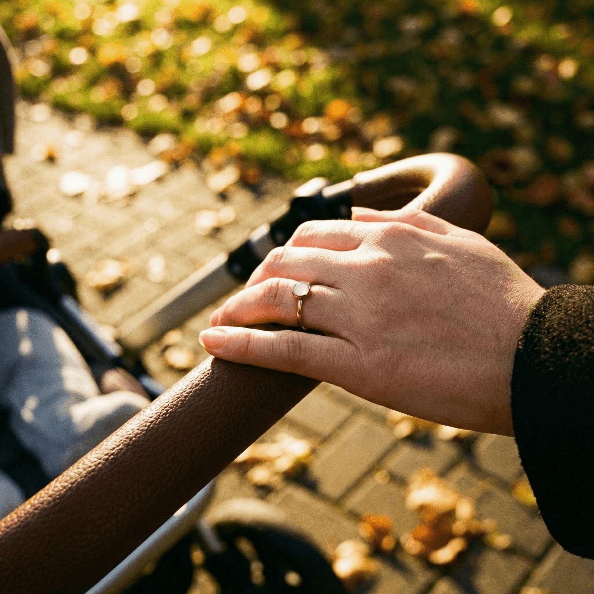 Mother’s hand holding a stroller handle outdoors in warm sunlight, wearing a delicate gold breastmilk ring with a small milky-white stone.