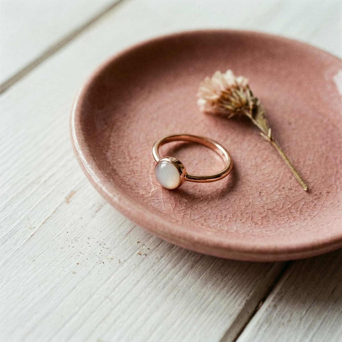 Rose-gold keepsake ring with a milky white stone on a blush ceramic dish beside a dried flower, showing the result of a DIY breastmilk ring kit.