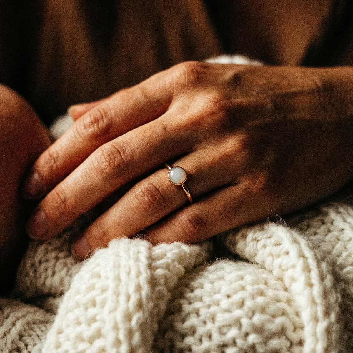 Mother’s hand wearing a minimalist gold ring with an opaque white stone keepsake made using a breastmilk ring making kit, resting on a soft knitted blanket.
