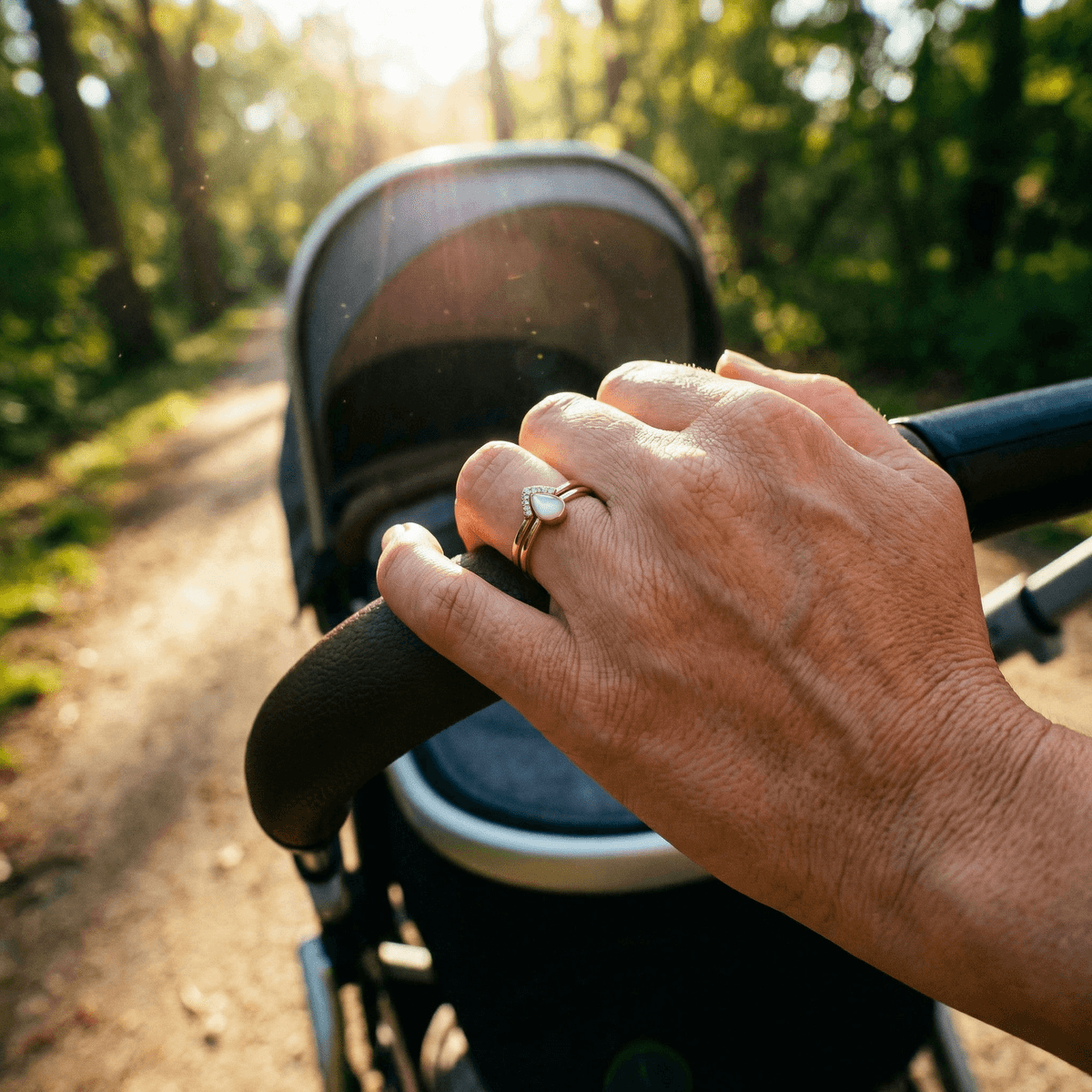 Mother outdoors pushing a stroller in warm sunlight, wearing elegant breastmilk rings featuring a teardrop milk-stone set in gold with pavé accents.