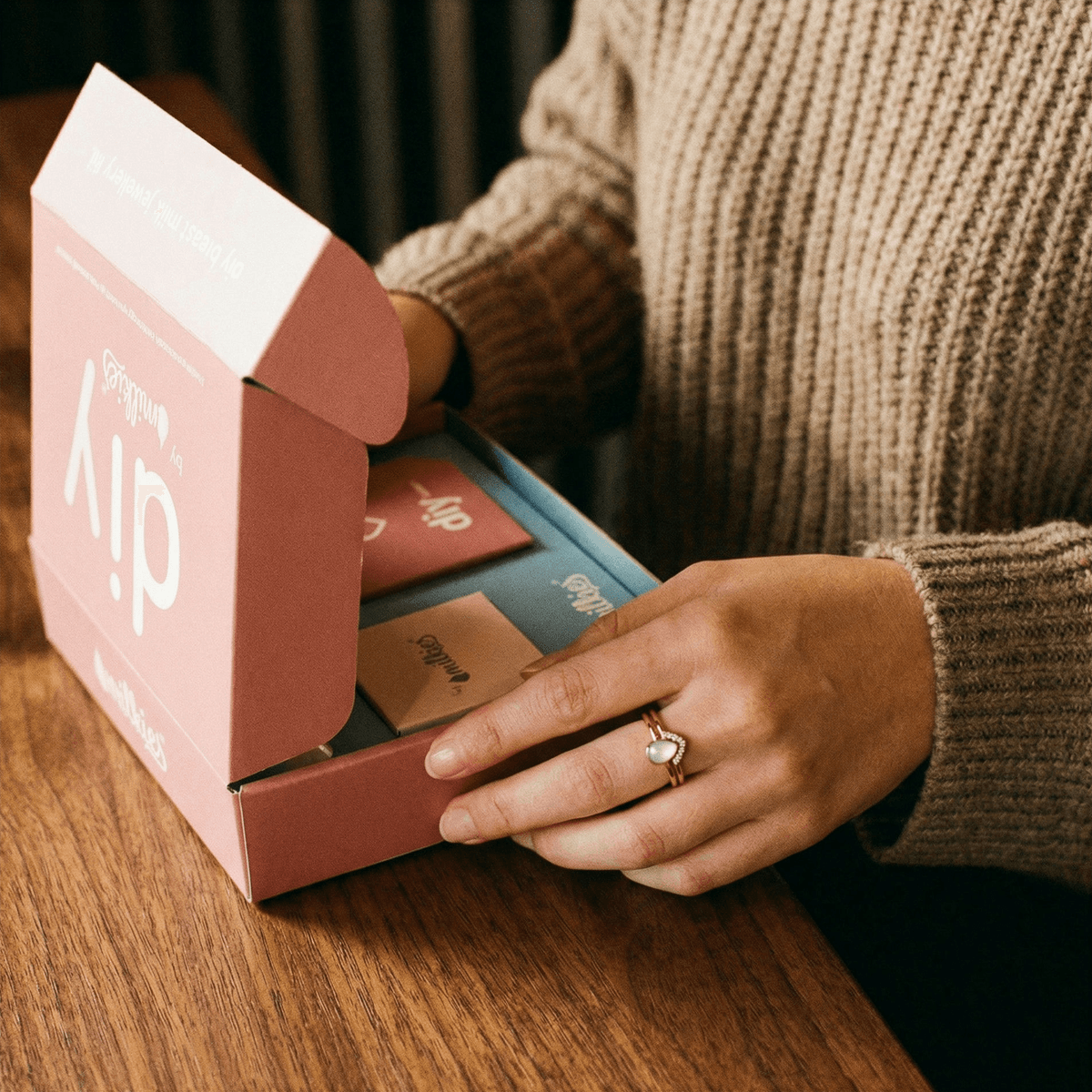 Hands open a DIY by MILKIES at-home keepsake kit on a wooden table, showing the guided, private process mothers use to make breastmilk rings and other jewelry at home.