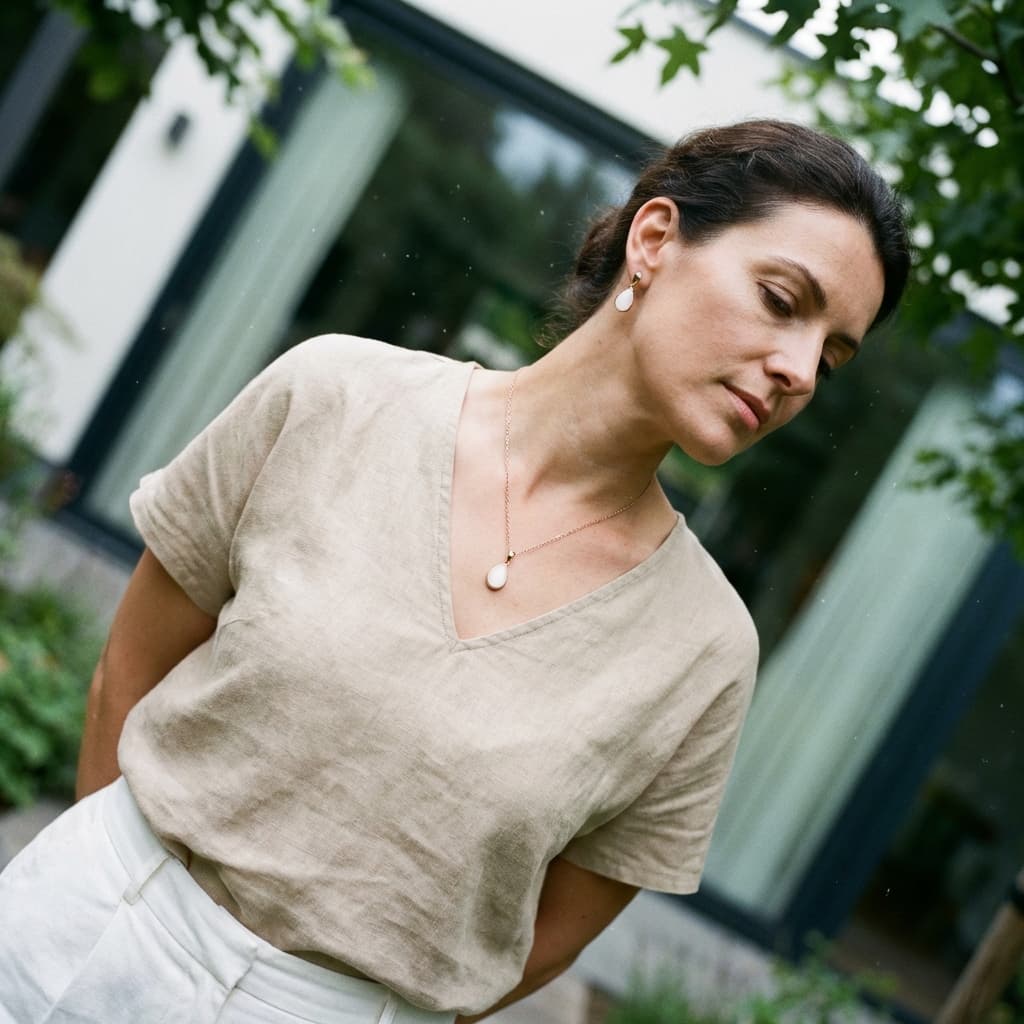 Woman outdoors wearing a minimal diy breast milk jewellery set with pearl-like pendant necklace and matching drop earrings, styled with a beige top.