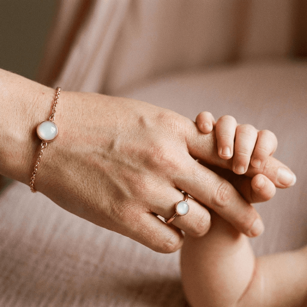 Close-up of a mother holding a baby’s hand while wearing a delicate diy breastmilk bracelet with a milky white resin stone in a rose-gold setting, paired with a matching ring.