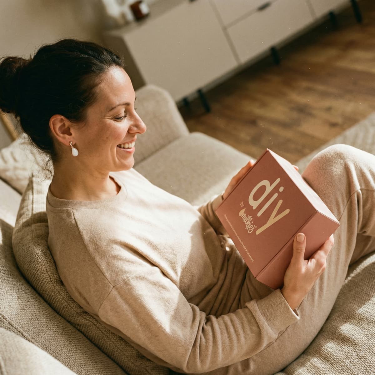 Smiling mom relaxing on a sofa holding a DIY by MILKIES kit box for making a diy breastmilk earring keepsake at home, highlighting the easy, private hands-on option.
