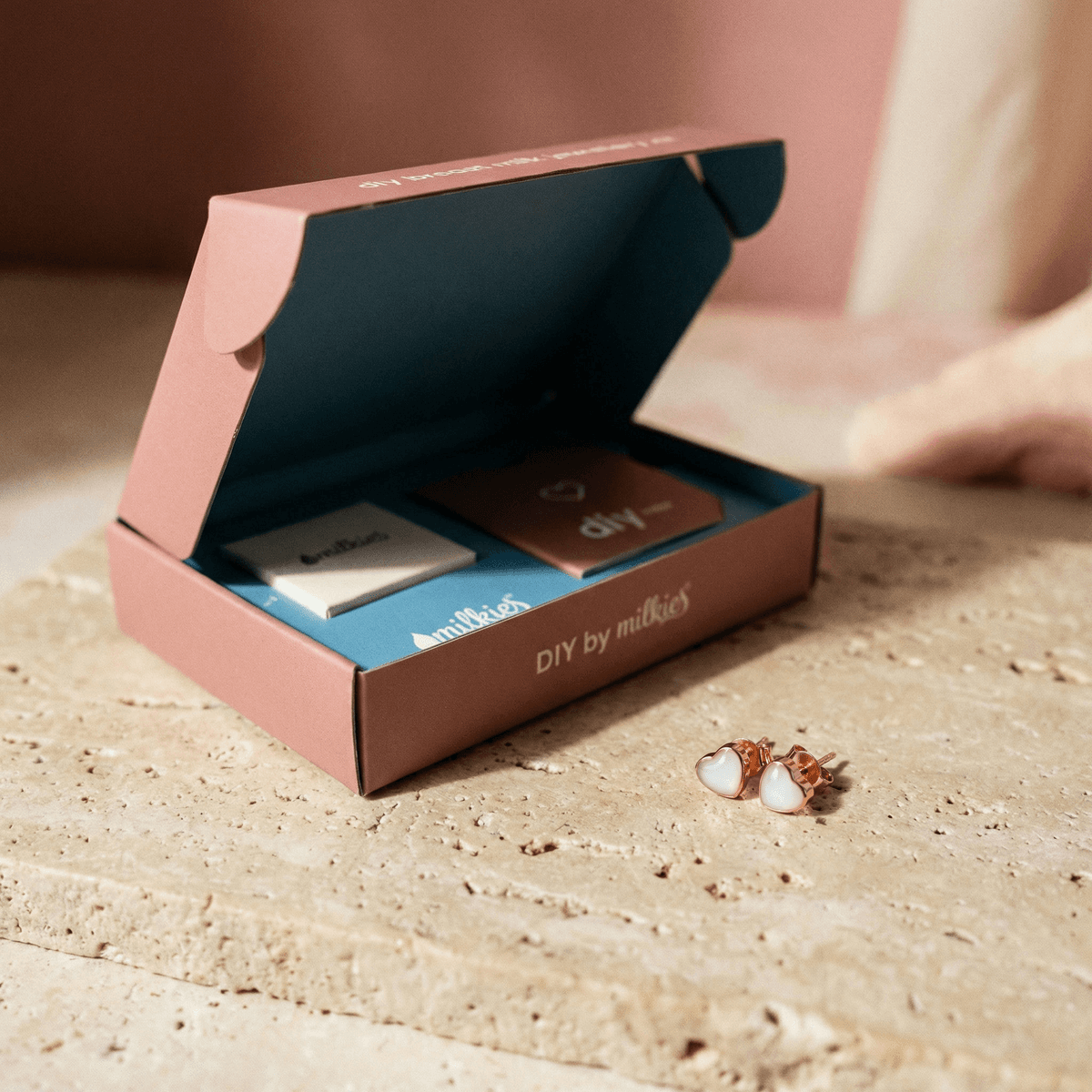 Open DIY by MILKIES box showing materials and rose-gold heart stud earrings on a stone surface, featuring an at-home diy breastmilk earring kit for creating keepsake jewelry.