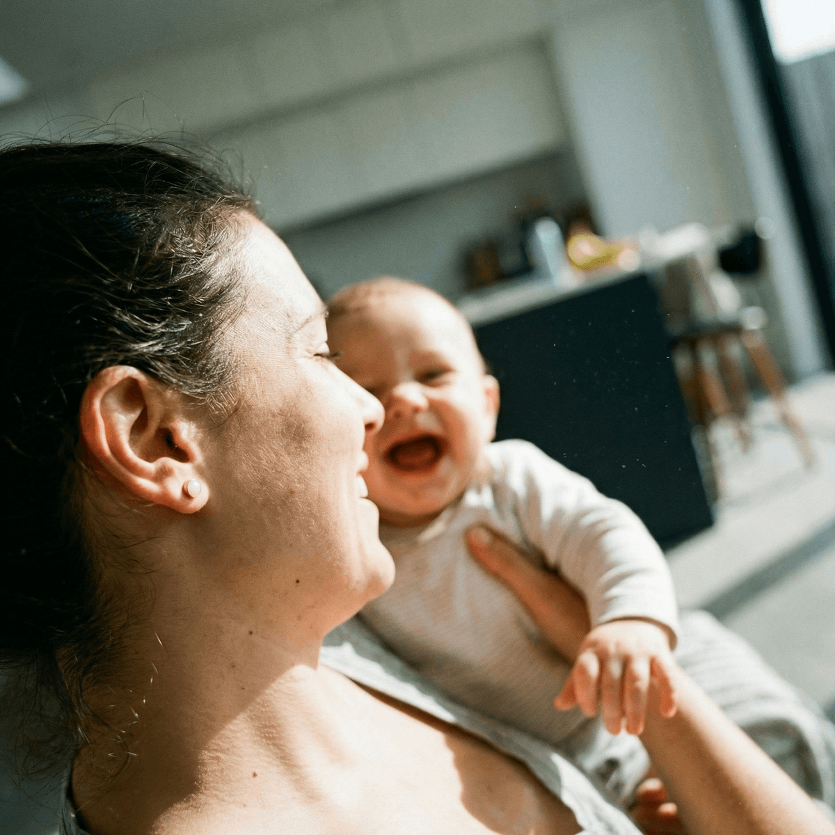 Mother holding a laughing baby in a sunlit home, wearing a stud earring that represents a diy breastmilk earring kit keepsake.