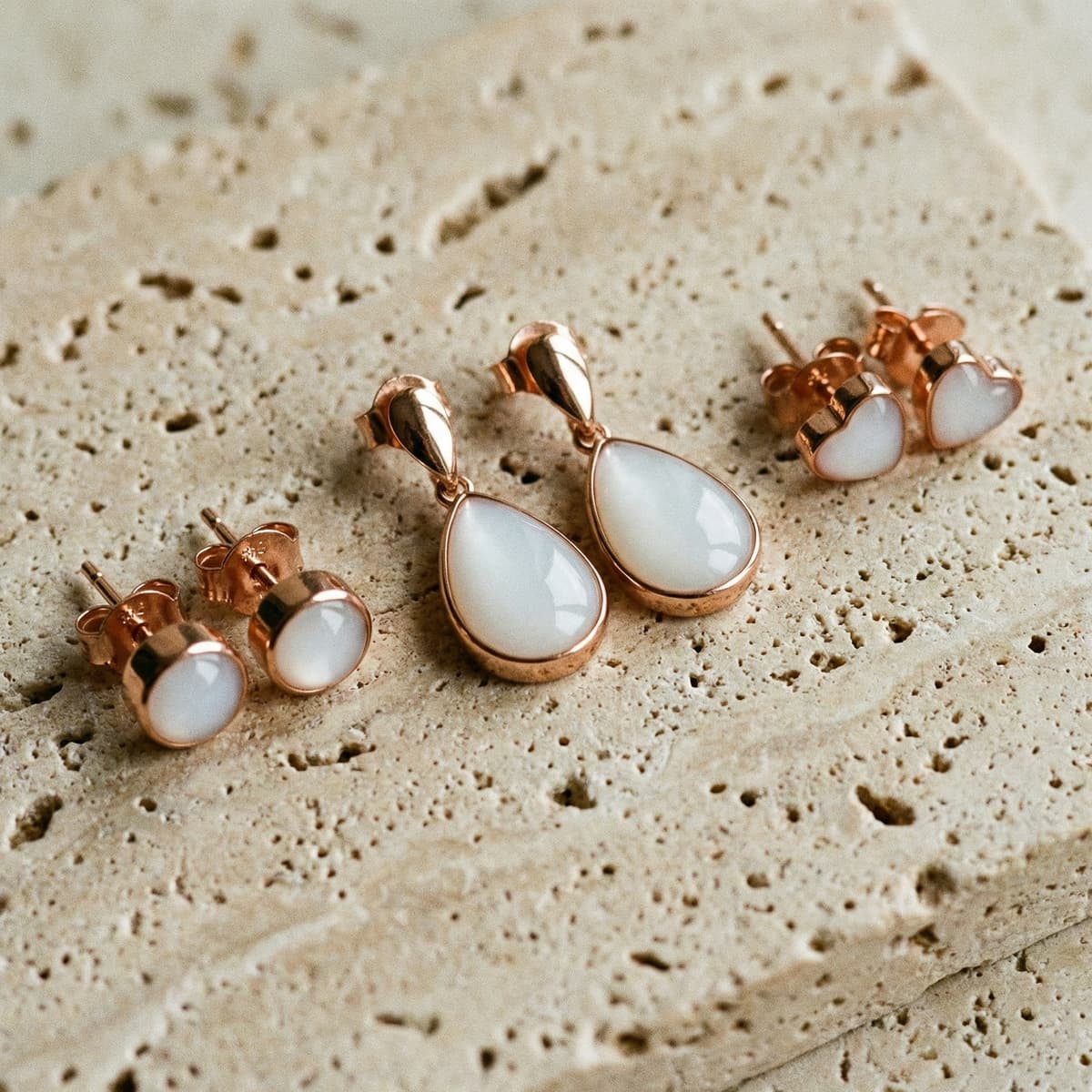 Rose gold plated teardrop and heart stud diy breastmilk earrings with milky white resin stones on a textured beige stone surface