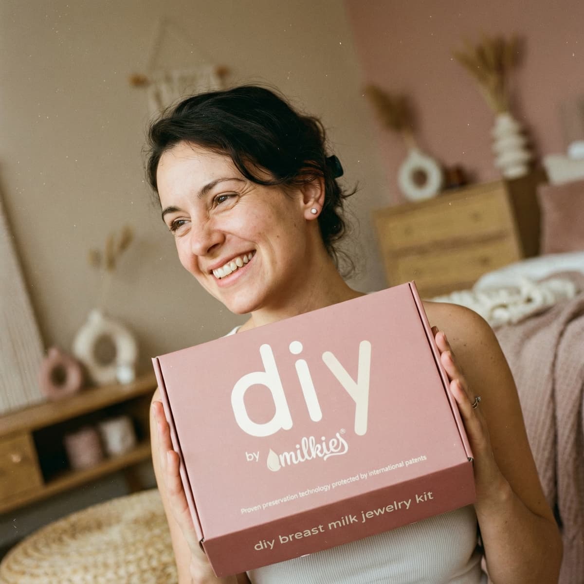Smiling mom holding a pink DIY by MILKIES breastmilk jewelry kit box, showing why DIY by MILKIES is an easy at-home choice for making diy breastmilk earrings with patented preservation technology.