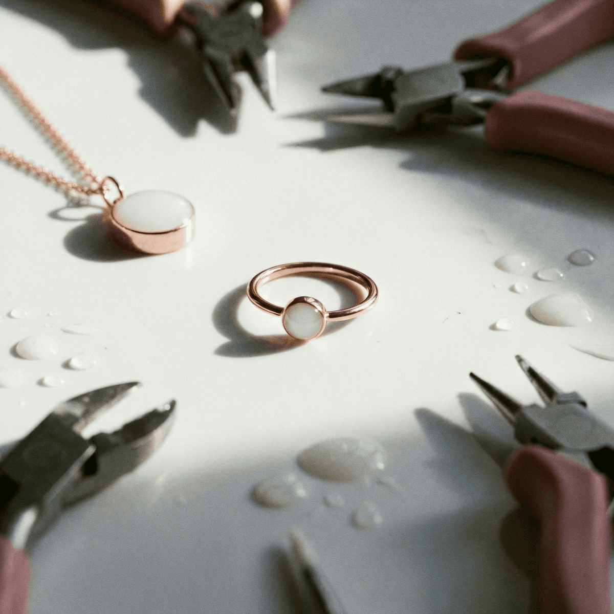 Close-up of a rose-gold ring kit featuring a milky resin stone, showing a diy breastmilk ring in progress on a white work surface with jewelry pliers, a matching pendant, and small drops of liquid.
