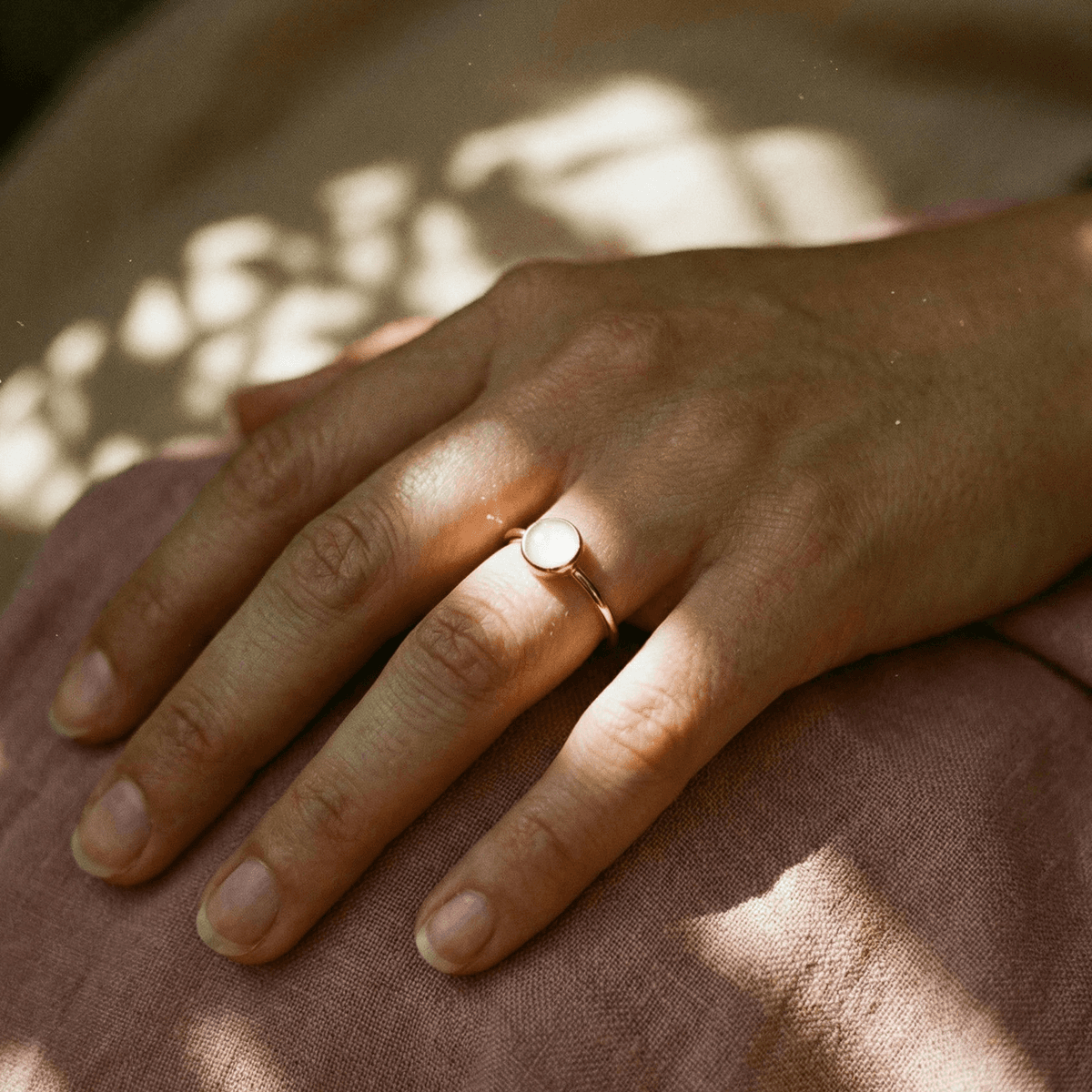 Hand wearing a delicate rose-gold DIY breastmilk ring with a round milky-white resin stone, resting on soft pink fabric in warm natural light.