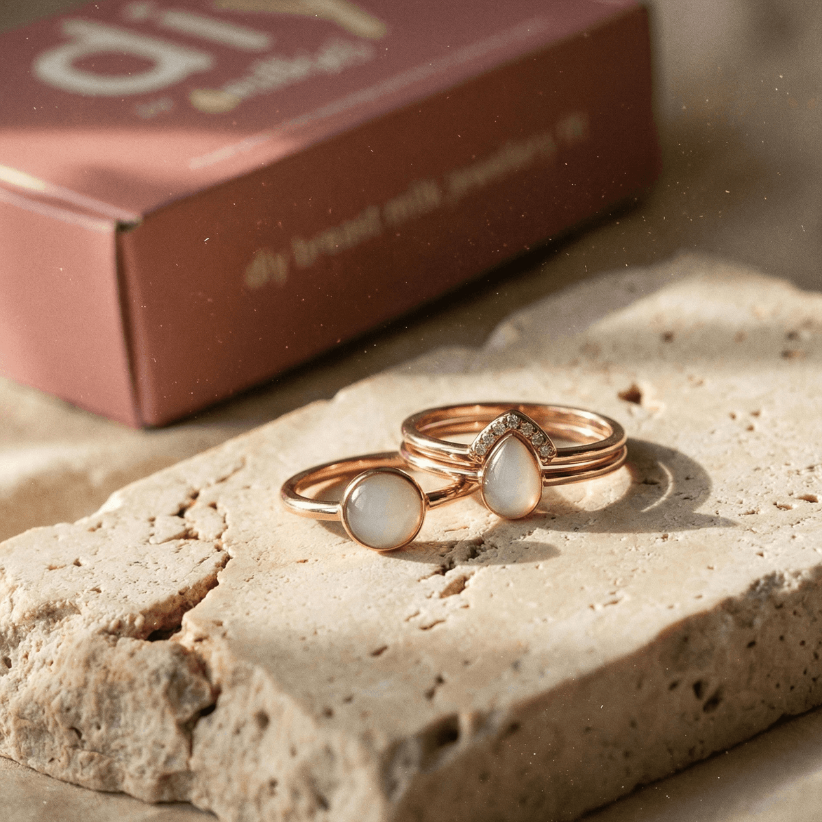 Rose gold rings with milky white resin stones on a textured stone slab beside a branded box, showcasing a diy breastmilk ring kit keepsake jewelry set.