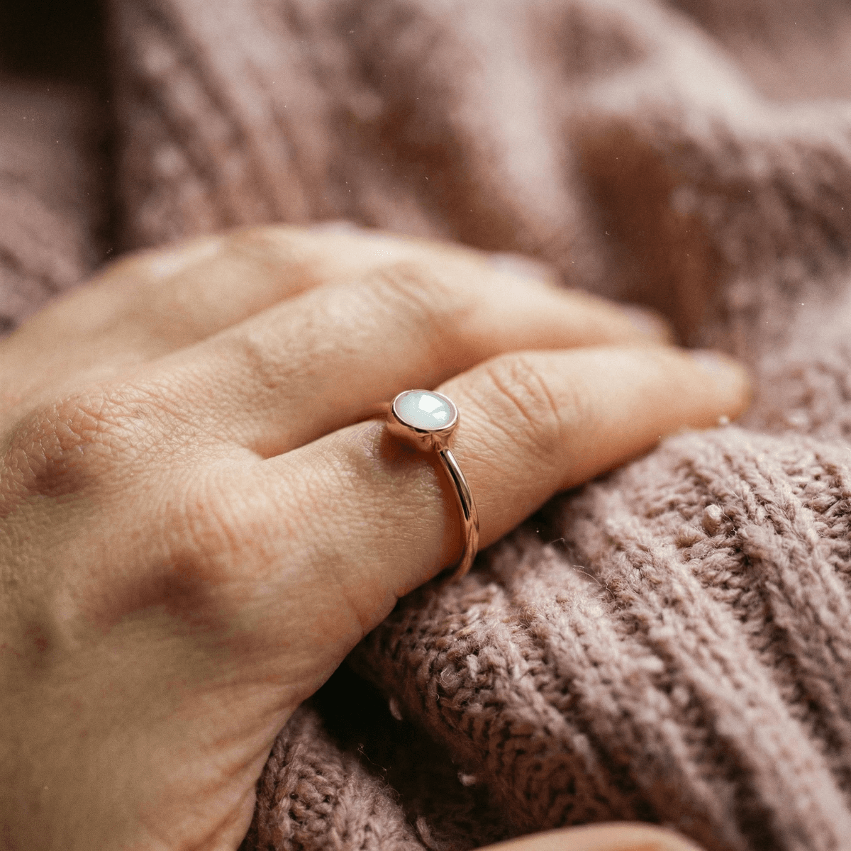 Close-up of a hand wearing a rose gold ring with a milky white resin stone, showcasing diy breastmilk rings as a personalized keepsake on a soft knitted blanket.