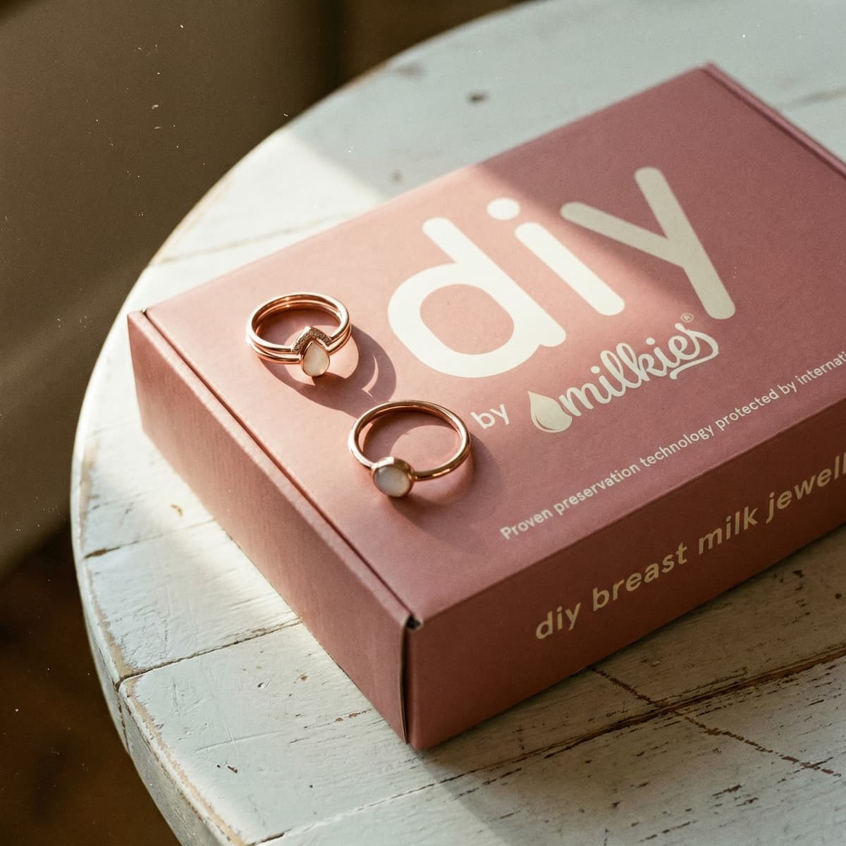 Two rose-gold DIY by Milkies kits featuring diy breastmilk rings with milky white stones on a pink kit box, styled on a rustic white table in warm natural light.