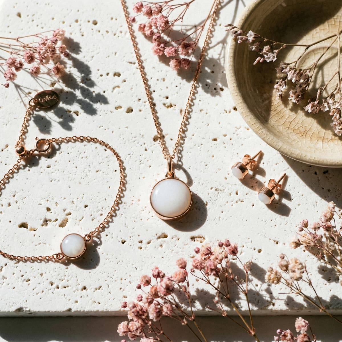 Rose-gold necklace pendant, stud earrings, and bracelet with milky white resin stones arranged on a textured stone surface with pink dried flowers and a ceramic dish, a delicate end of breastfeeding gift jewelry set.