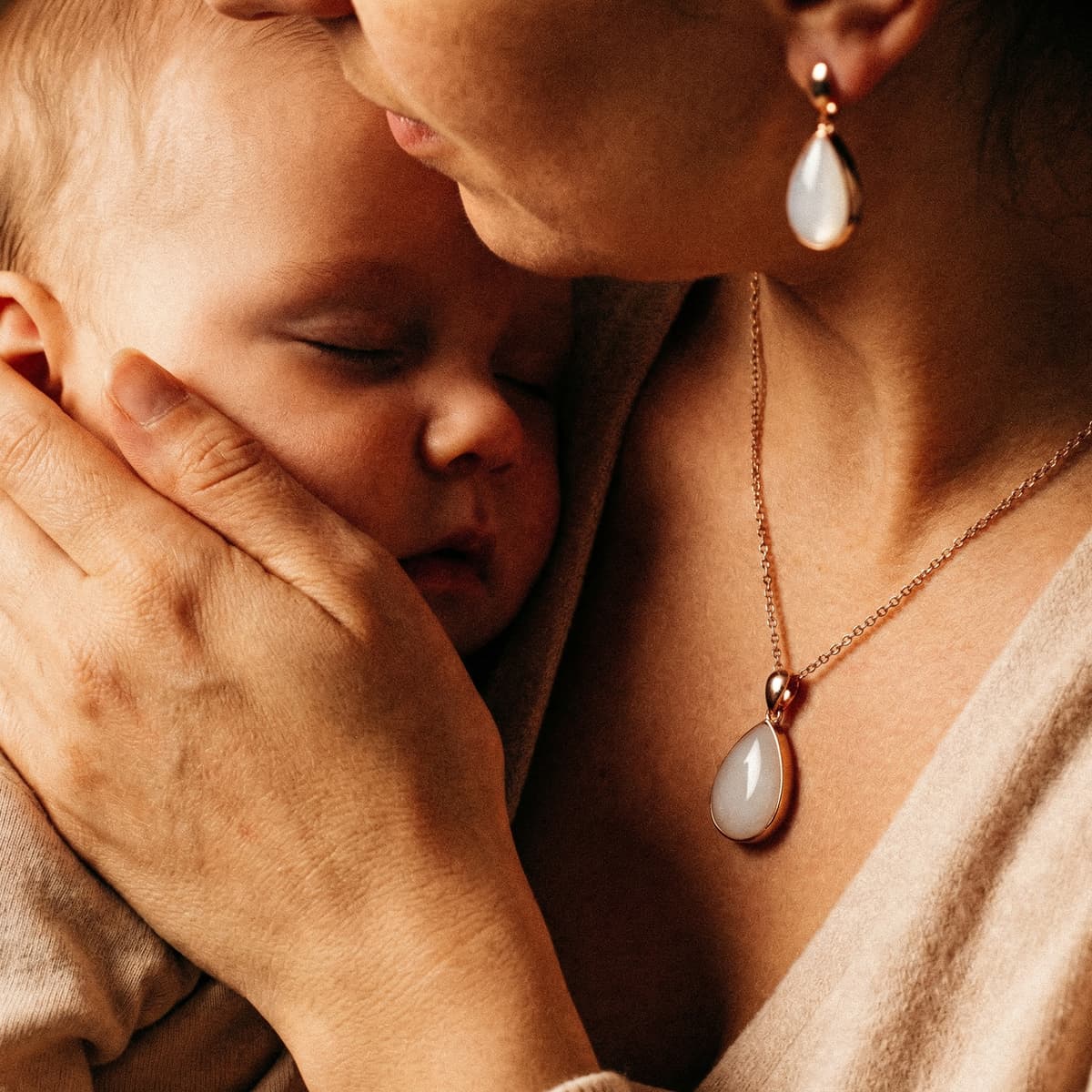 Mother cuddling a sleeping baby while wearing delicate breastmilk keepsake jewelry inspired by golden boobs, featuring a white teardrop pendant necklace with matching earrings.