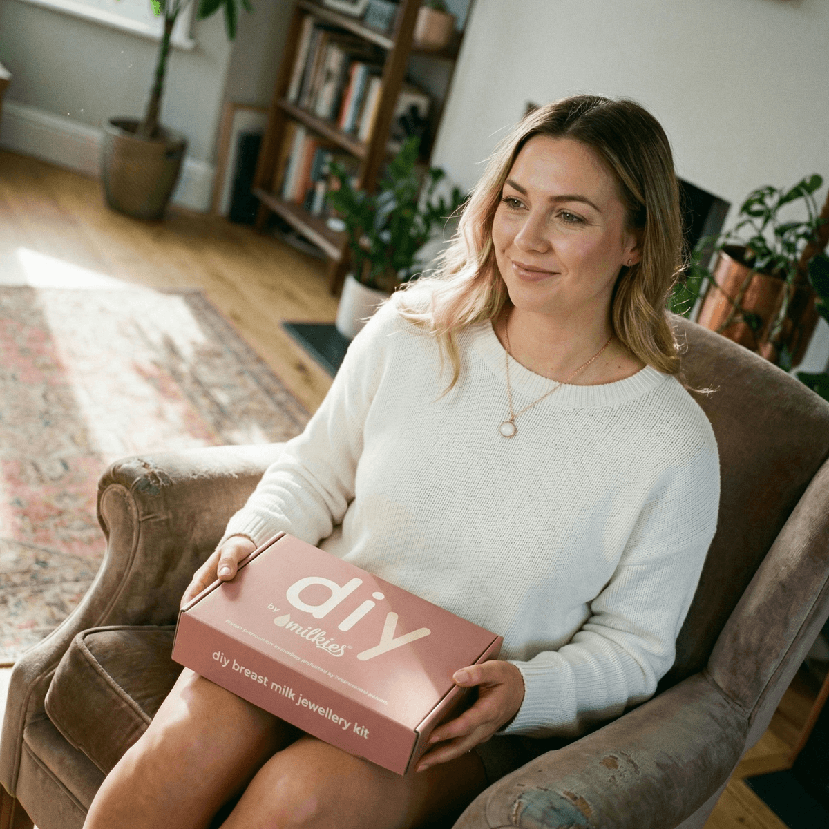Mom in a cozy living room holding a pink DIY by MILKIES breast milk jewelry kit box, highlighting why the at-home kit is a convenient option for creating a last latch keepsake in privacy.