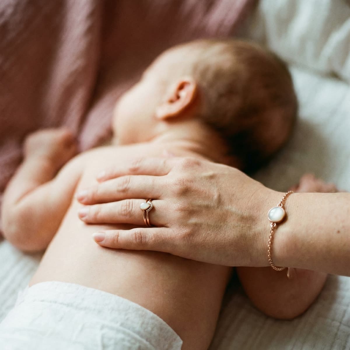 Mother cradling a sleeping baby while wearing a rose-gold ring and bracelet with milky white stones, a nursing keepsake jewelry set.