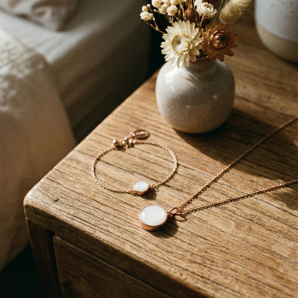 Rose-gold breastmilk keepsake necklace and bracelet with milky white resin stones displayed as an elegant push present on a rustic wooden bedside table beside a small vase of dried flowers.