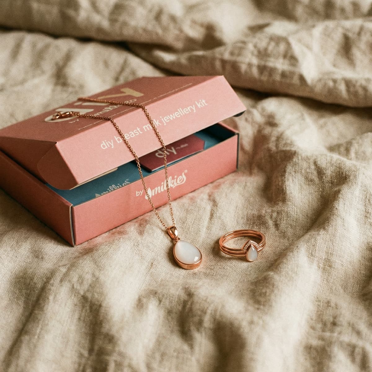 DIY by MILKIES breast milk jewellery kit box with rose-gold necklace pendant and matching ring on a soft bedspread, a meaningful push present for wife to create a breastmilk keepsake at home