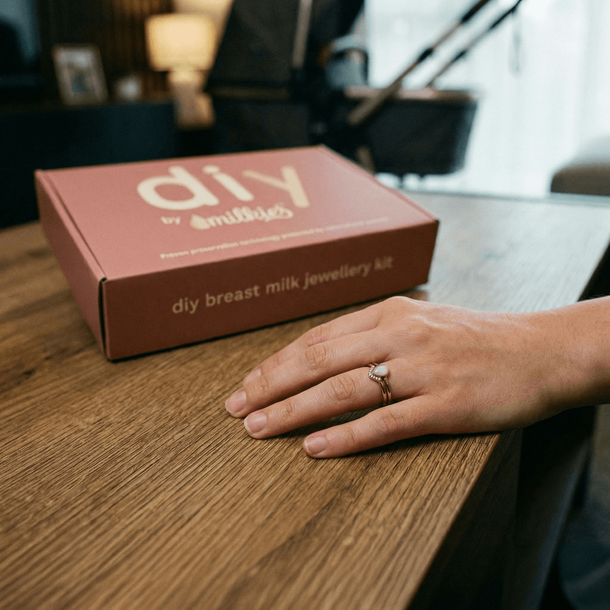 Hand resting on a wooden table wearing a breastmilk keepsake ring next to a DIY by MILKIES breast milk jewellery kit box, showcasing a thoughtful idea for unique push presents.