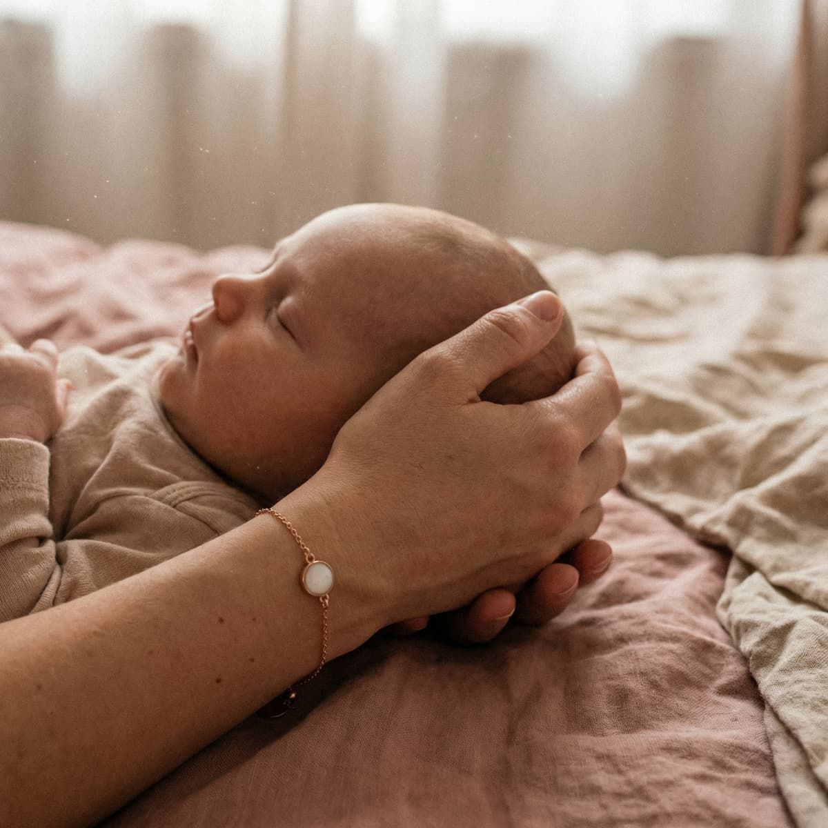 Mother cradling a sleeping newborn on a bed while wearing a keepsake bracelet, highlighting why a breastmilk bracelet diy making kit is a meaningful at-home way to create a personal breastmilk jewelry memory.