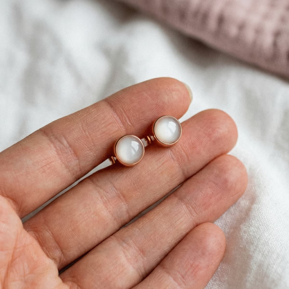 Close-up of a hand holding two round rose-gold stud earrings with milky white resin stones, a finished keepsake made using a breastmilk earring diy making kit.