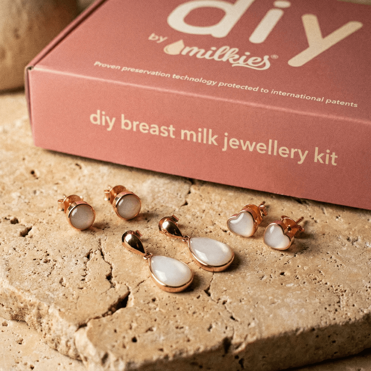 Rose-gold breastmilk earrings displayed with heart studs, round studs, and teardrop dangle design in front of a breastmilk earrings DIY making kit box by DIY by MILKIES.
